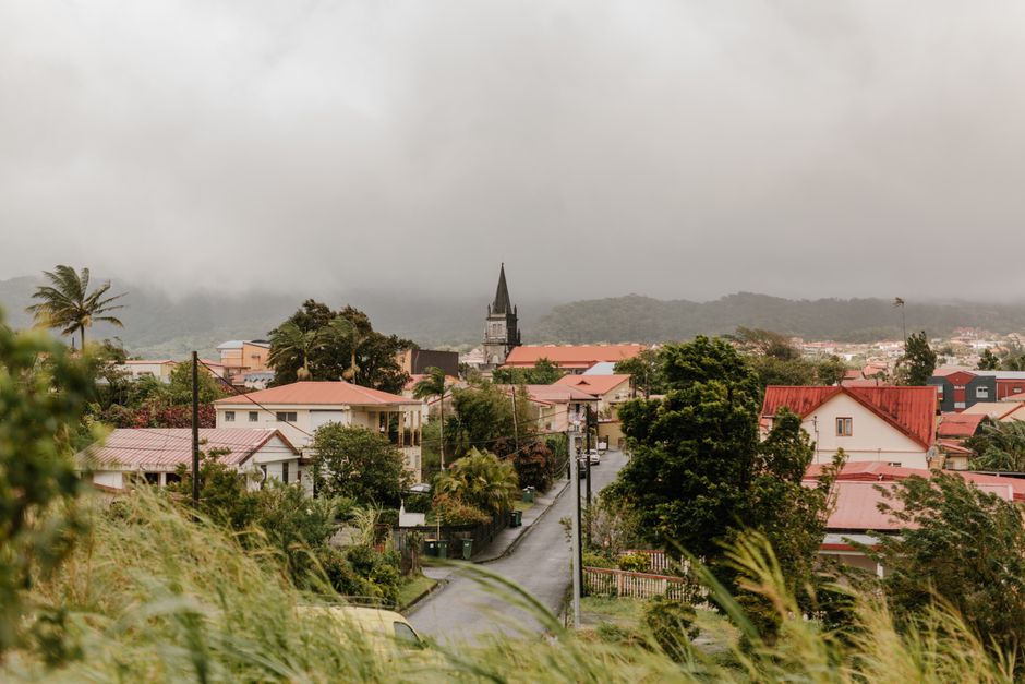 A view of a residential area in De Beauvoir Town featuring a narrow paved street lined with various houses, some with red tiled roofs and others with light-colored exteriors. In the foreground, there is lush greenery and tall grass on an incline, partially obscuring the view. The street appears to be part of a home relocation or moving process, as a man with a van from Man with Van De Beauvoir Town may be involved in furniture transport or packing activities. A church steeple is visible in the distance, set against a backdrop of low-lying hills shrouded in overcast, cloudy weather, creating a subdued atmosphere suitable for highlighting moving and packing logistics within a typical suburban neighborhood.