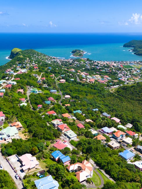 Aerial view of a residential area with numerous houses and greenery extending towards a coastline with a blue sea and small islands visible in the distance. The houses are various colors, with some roofs in shades of red, blue, and brown. The area appears densely populated with tree-lined streets and lush vegetation surrounding the homes. The coastline features sandy beaches and calm waters, under a partly cloudy sky. This scenic landscape represents a coastal community, ideal for house removals and relocation services, with the natural environment providing a peaceful backdrop for moving logistics managed by Man with Van De Beauvoir Town.