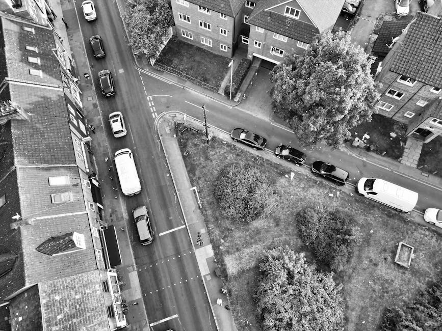 Black and white aerial photograph of a residential street scene showing a line of parked cars along the curb on the right side of the street, with some angled towards the pavement and others parallel. The street is narrow, with a curve leading to a small corner, and has road markings including a pedestrian crossing. On the left side, a row of terraced houses with tile roofs and front entrances is visible. In the foreground, a white van is parked just off the street. Nearby, a small grassy area contains two large trees and some bushes, with a narrow pathway separating the greenery from the pavement. The scene captures the environment in which house removals or furniture transport might take place, with parked vehicles and a typical residential setting. This image aligns with the relocation and house moving services offered by Man with Van De Beauvoir Town, highlighting urban loading areas suitable for home relocation and packing and moving processes.