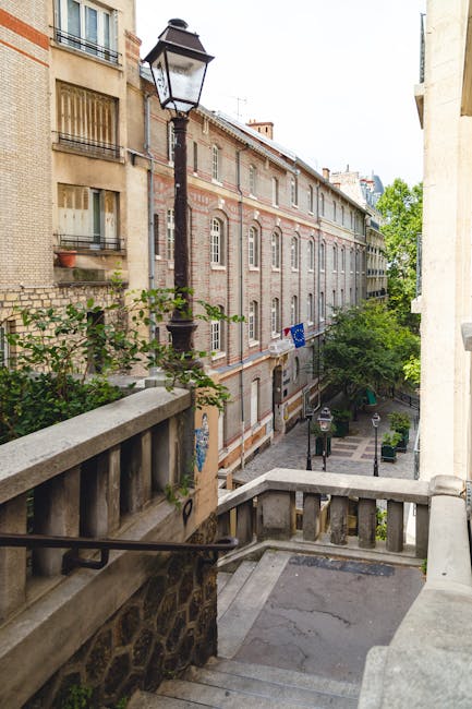 A view from a balcony overlooking a narrow street in a residential area, featuring multi-storey brick and stone buildings with large windows and balconies, a vintage-style lamppost located on the sidewalk, and a small green tree near the edge of the street. The scene shows part of the staircase leading down from the balcony to the pavement, with a stone balustrade and decorative railing. The street is lined with parked vehicles, and there are signs of greenery, such as potted plants and trees, indicating an urban environment typical for house removals in De Beauvoir Town. This setting provides context for the logistics involved in home relocation, highlighting the urban architecture and street layout where furniture transport and packing and moving processes are managed by companies like Man with Van De Beauvoir Town.