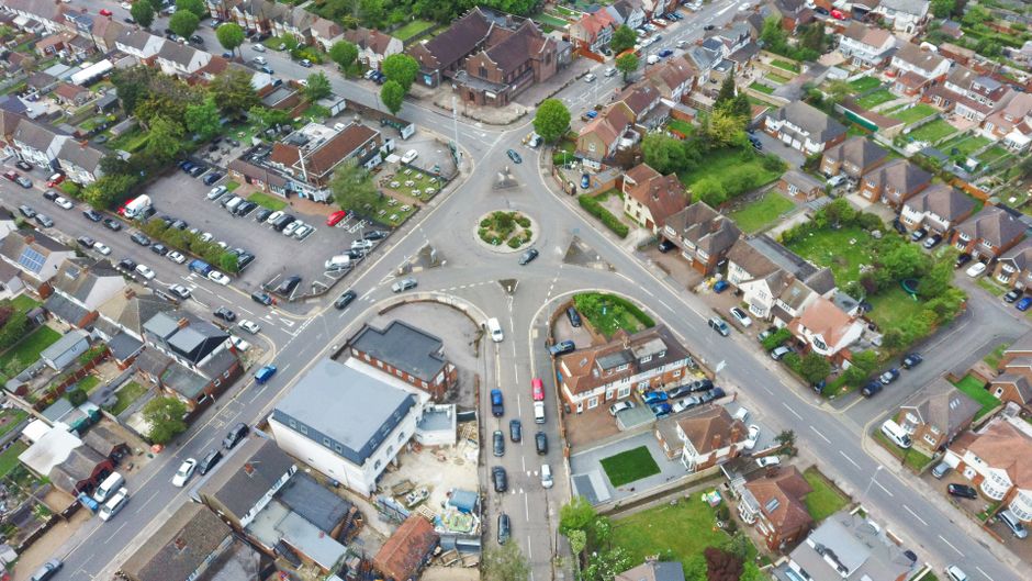 An aerial view of a residential urban area showing a roundabout intersection surrounded by houses, gardens, and parked cars. The roundabout includes a small landscaped island with greenery and a circular central feature. Multiple roads branch from the roundabout, with cars parked along the streets and some in motion. In the lower left corner, an area under construction is visible, with a partially built structure, construction materials, and equipment. The image captures the street layout and housing density characteristic of a typical neighbourhood, with trees and green spaces interspersed among the houses. This scene relates to house relocations and transport logistics, similar to services offered by Man with Van De Beauvoir Town, highlighting urban moving and packing environments, including loading and unloading points in residential areas.