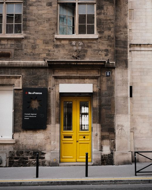 The image features the exterior facade of a building in De Beauvoir Town, with a prominent bright yellow door centered in the photograph. The door has two vertical glass panes with black framing and a small, round door handle on the right side. To the left of the door, there is a black sign with a gold star-shaped emblem and white text indicating the location as the regional council's office for Iliad France, attached to a weathered stone wall with visible age marks and textures. Above the door, there is a white, rectangular signboard, possibly for posting notices, and the building's facade includes three windows with old, worn frames—two situated directly above the door and one to the left, all with glass panes reflecting ambient light. The steps of the sidewalk are paved with light grey concrete, with black safety bollards along the edge, and a low black metal railing on the right side of the frame. The scene appears to be lit with natural daylight, capturing a neutral, urban environment suitable for home relocation or moving services in the area, as illustrated by the building's character and the typical street scene outside a residential or small commercial property.
