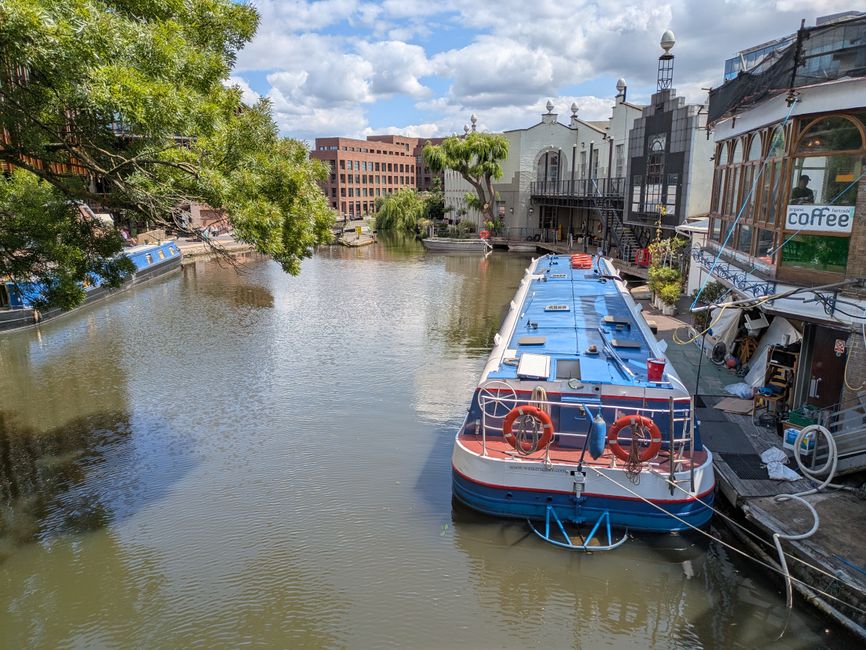 A narrowboat moored along the towpath of Regents Canal during daytime, with its blue and white exterior visible. The boat is tied securely to the bank with ropes, and various equipment such as life rings and fenders are attached to the stern. Adjacent to the boat, there is a small outdoor area with a building that has large windows, some with visible signage including a coffee shop. The scene shows a partly cloudy sky, with trees and modern buildings in the background. The towpath is clear, and the waterfront setting is suitable for home relocation or furniture transport activities, as managed by Man with Van De Beauvoir Town, highlighting typical logistics involved in packing and moving operations along canal routes.