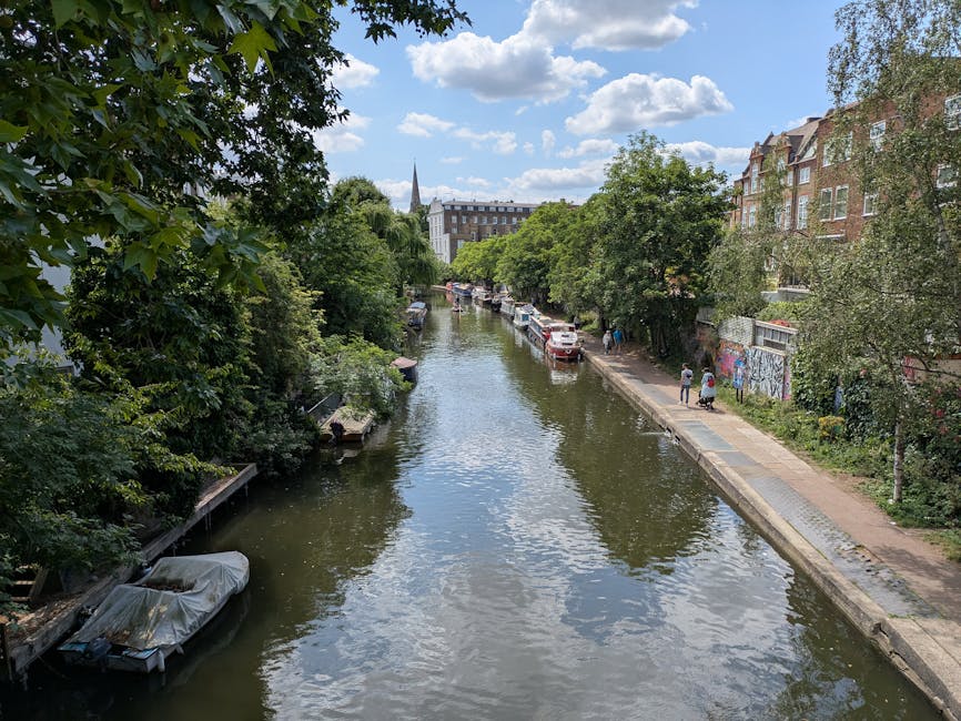 A narrow canal lined with lush green trees on both sides, with several boats moored along the edges, one covered with a white tarp. On the right side, a paved footpath runs parallel to the canal, with a few pedestrians walking. Parked cars are visible along the street behind the trees, and multi-story residential buildings with brick facades and large windows border the right side of the scene. In the distance, a church spire rises above the treetops, and the sky is partly cloudy with blue patches. The overall setting reflects an urban residential area suitable for house removals and furniture transport, with nearby waterway access and careful consideration needed for loading and unloading process during home relocation, as seen in the context of delivery issues along the Regents Canal, with visible moving equipment, such as a trolley or straps, on the pavement nearby.