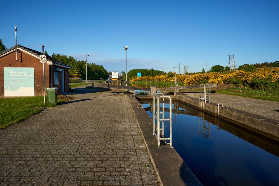 A narrowboat moored along the towpath of Regents Canal during daytime, with its blue and white exterior visible. The boat is tied securely to the bank with ropes, and various equipment such as life rings and fenders are attached to the stern. Adjacent to the boat, there is a small outdoor area with a building that has large windows, some with visible signage including a coffee shop. The scene shows a partly cloudy sky, with trees and modern buildings in the background. The towpath is clear, and the waterfront setting is suitable for home relocation or furniture transport activities, as managed by Man with Van De Beauvoir Town, highlighting typical logistics involved in packing and moving operations along canal routes.
