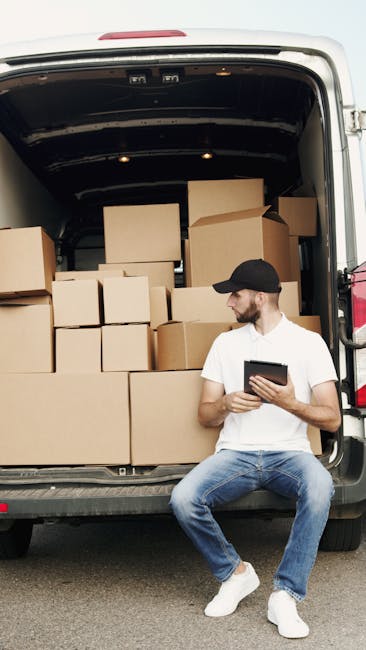 A man wearing a white t-shirt and jeans stands outside in daylight, preparing to load cardboard boxes into the open rear cargo area of a white van. The boxes are stacked on the pavement beside him, so