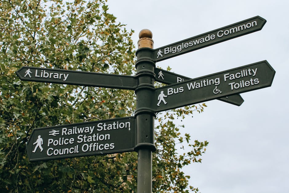 A black multi-directional signpost situated outdoors against a background of green trees and an overcast sky, featuring several white directional arrows with labels indicating nearby locations. The arrows point towards Biggleswade Common, a library, a bus waiting facility and toilets, a railway station, police station, and council offices. The signpost is secured to a metal pole, with each arrow displaying walking icons and relevant symbols for accessibility, such as a wheelchair. This scene is related to urban navigation and community infrastructure, relevant to the context of home relocation or moving services provided by Man with Van De Beauvoir Town, highlighting the importance of understanding local amenities and directional guidance involved in furniture transport and packing during house removals.