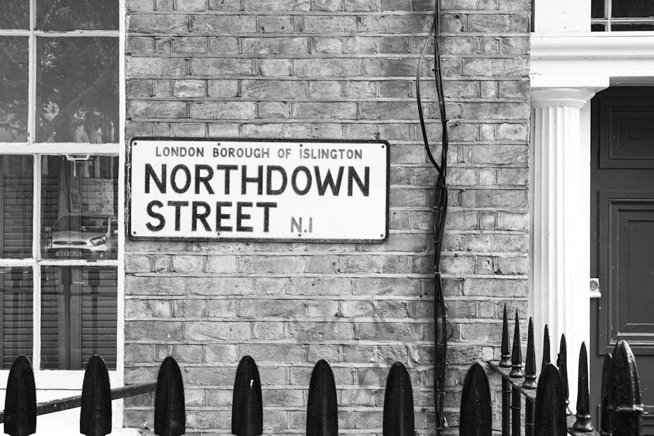 A black and white photograph of a residential building facade on Northdown Street, Islington, showing a brick wall with a street sign that reads 'London Borough of Islington Northdown Street N.1'. To the left, there is a window with multiple panes, reflecting parked cars outside. Visible are several cardboard boxes and packing materials stacked near the doorway, ready for home relocation or furniture transport. A dark-colored entrance door with molding details is partially visible on the right side. In the foreground, a low metal fence with pointed finials separates the property from the pavement. A cable or wire is loosely draped along the wall, indicating preparation for moving or electrical work. The scene depicts the preparatory stage of a house removal, highlighting the importance of organized packing and access for loading furniture and boxes onto the van, supported by a moving company such as Man with Van De Beauvoir Town familiar with local council regulations affecting such procedures.