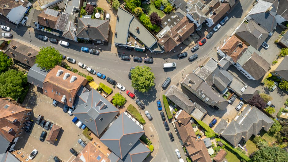 An aerial view of a residential neighbourhood showing a narrow, curved street lined with parked cars on both sides, with houses featuring pitched roofs, some with chimneys and dormer windows, and small front gardens with trees and hedges. In the foreground, a driveway with a grey roofed house has a collection of cardboard boxes, wrapped furniture, and packing materials indicating an ongoing home relocation process. Nearby, a man with a van marked 'Man with Van De Beauvoir Town' is seen loading or unloading large cardboard boxes and furniture items onto or from a van parked on the street, using tools such as trolleys or straps for safe handling. The surrounding area includes detached and semi-detached houses with various building materials like brick and weatherboard, and the overall scene captures the logistics of furniture transport and packing during a move within the local area, highlighting careful loading and organized movement typical of professional removals services.