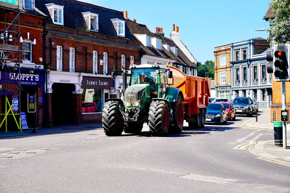 A large green tractor with an orange trailer attached is positioned at an intersection on a city street, surrounded by parked cars and historic brick buildings. The tractor appears to be involved in a home relocation or moving process, as part of the wider logistics of furniture transport. The street scene features a traffic light displaying red, and the weather is sunny with clear blue skies. The buildings include retail shops such as a sports bar and a salon, and their facades display large windows with decorative elements. The tractor's size and design indicate it is used for heavy-duty transport or loading activities, consistent with professional removals services like those offered by Man with Van De Beauvoir Town. The environment suggests an urban area with narrow streets conducive to moving and packing operations, supporting an efficient home or office move in the area.
