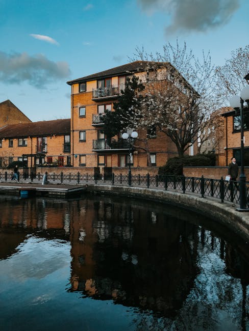 An exterior view of a multi-storey residential building with brick walls and balconies, situated beside a canal with dark, reflective water. The environment features a paved walkway with a black metal