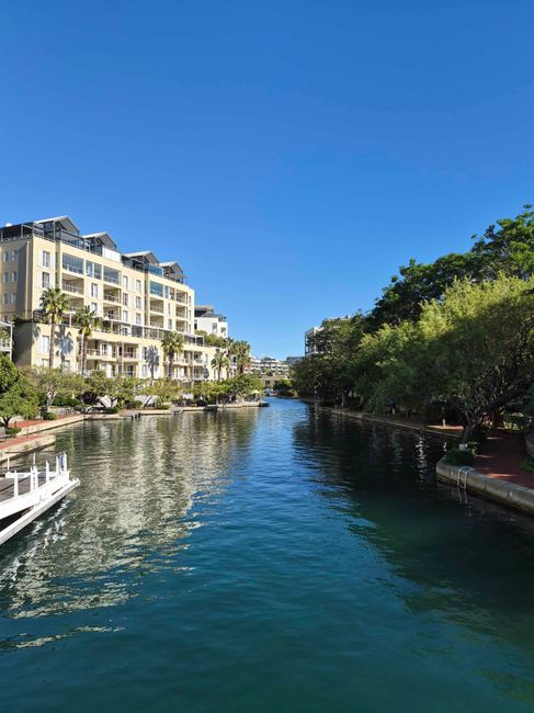 An exterior view of a multi-storey residential building with brick walls and balconies, situated beside a canal with dark, reflective water. The environment features a paved walkway with a black metal