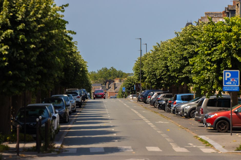 A quiet street scene in De Beauvoir Town featuring a parking and loading bay area. Several parked vehicles, including a black compact car and a vintage-style car with a spare tire mounted on the rear,