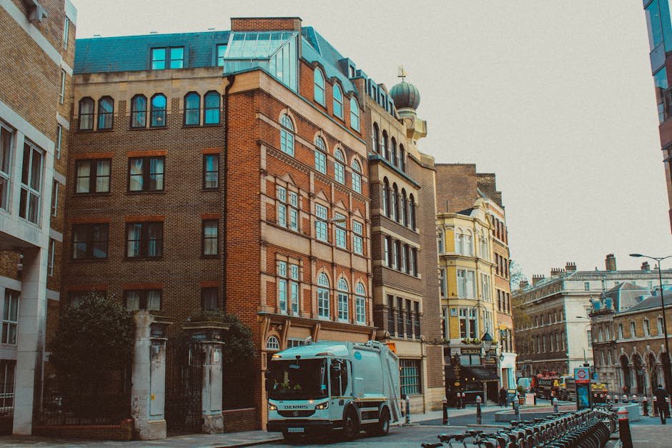 A light blue moving van parked on a busy urban street with old and modern buildings lining the pavement. The van is positioned near the curb with its side door closed, and there are several cardboard