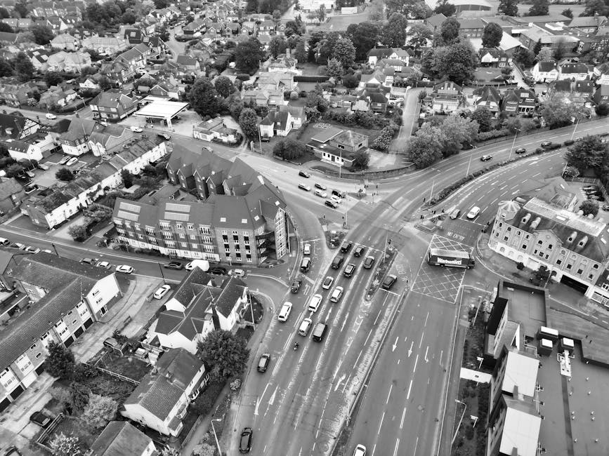 An aerial view of a residential neighbourhood showing a narrow street intersection with a roundabout, lined with terraced houses and semi-detached homes. Cars are parked along the road and in driveways, while some vehicles are in motion. The street runs through a lush, green area with trees and small gardens visible in front and behind the properties. The scene captures the typical layout of urban housing close to Southgate Road and Kingsland Road, relevant to home relocation and furniture transport services. The lighting suggests daylight, with natural light illuminating the rooftops, paving, and surrounding greenery. This setting illustrates the typical environment for a house removal or moving process, with visible residential details and street infrastructure.