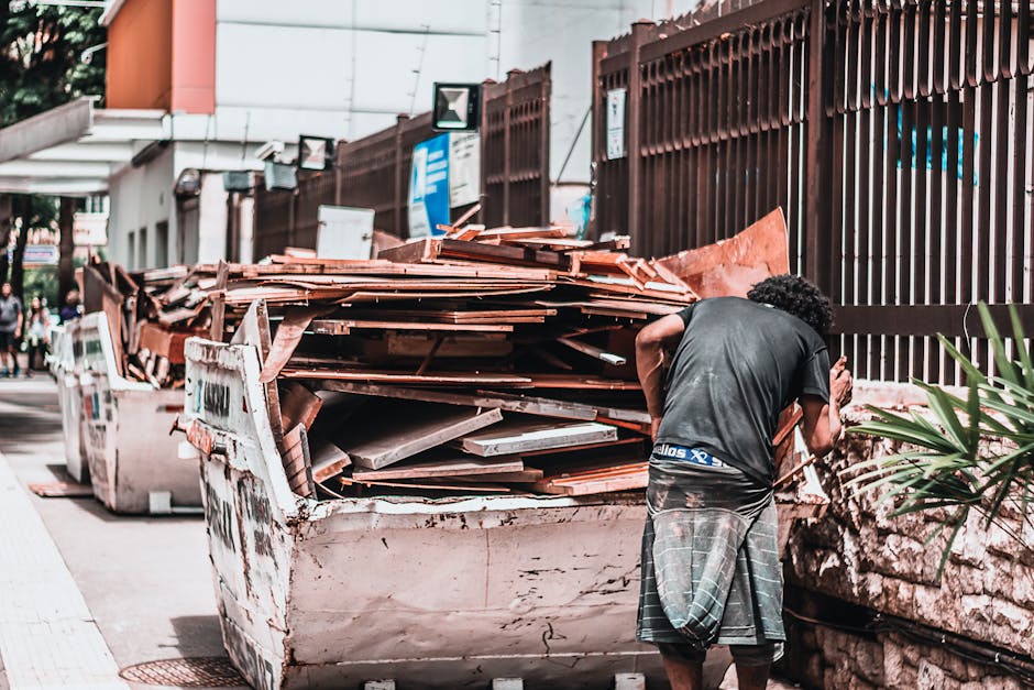 A person with curly dark hair, dressed in a dark t-shirt and checkered shorts, is positioned next to a large, overflowing skip on a city street during daytime, lifting or preparing to load bulky waste materials. The skip contains various discarded building materials, including wooden planks, ceramic tiles, and cardboard boxes, with some items stacked haphazardly. The waste is situated on the pavement adjacent to a stone wall and a metal fence. In the background, there are street signs, a building with a covered walkway, and a few pedestrians walking along the sidewalk. The scene, captured in natural daylight, depicts a typical home relocation or furniture transport process involving loading unwanted bulky items before a move, consistent with the services offered by Man with Van De Beauvoir Town, specialists in removals and moving logistics.